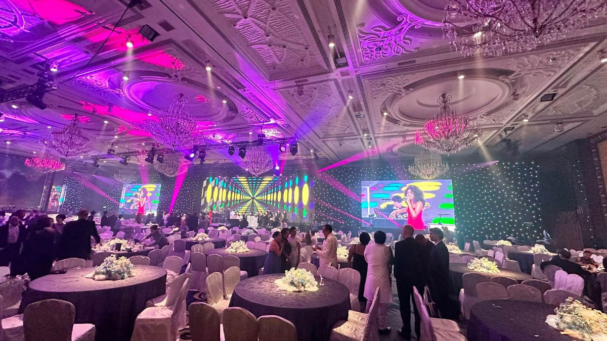 Guests stand between round tables under magenta and violet stage lighting in a hotel ballroom, with the orchestra on stage