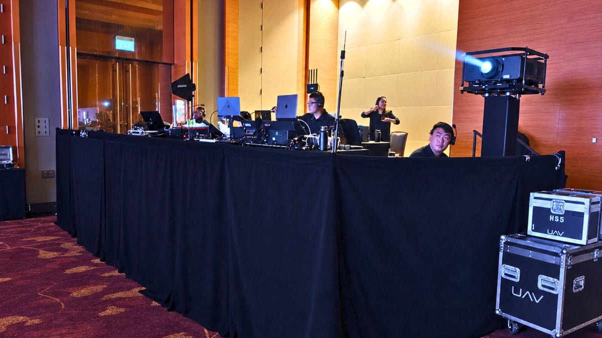 Crew in headsets at a black-draped control table in a function room, beside a projector on a stand and flight cases marked UAV