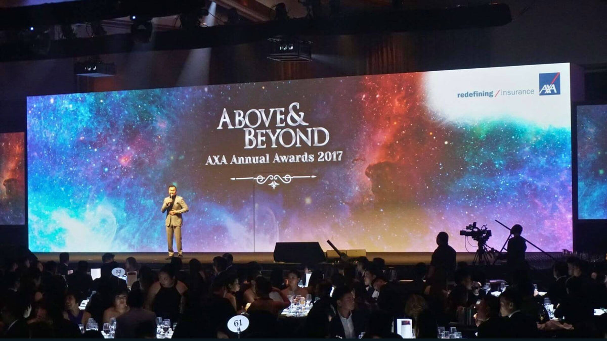 A presenter on stage in front of a wide LED wall at a gala dinner, with a camera operator at the side and guests seated at round tables.