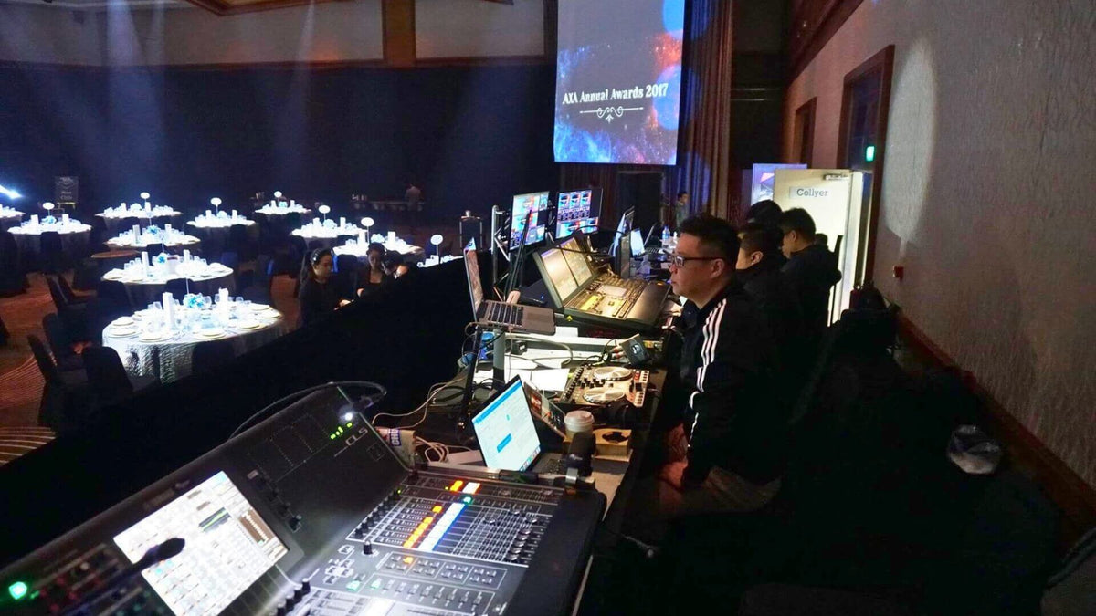Operators at a mixing console and lighting desk at the back of a ballroom, facing a projection screen above the dinner tables.