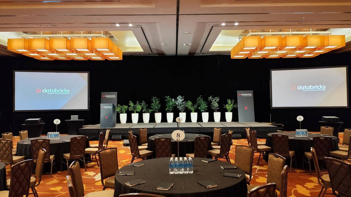 A ballroom set with round tables, two projection screens either side of a low stage, a lectern and a row of planters along the stage edge.