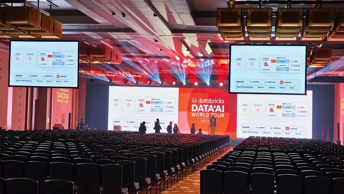 A conference room with a wide stage screen and two large repeat screens suspended either side, lit in red, with rows of chairs and people at the stage edge.