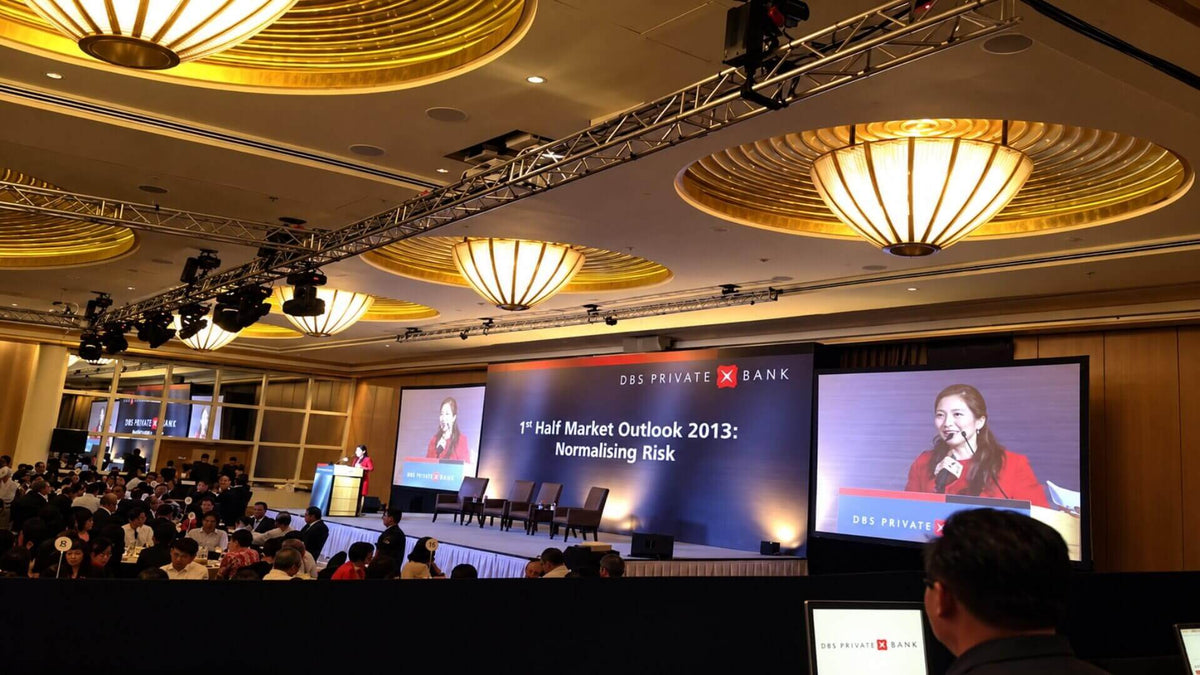 A conference in a hotel ballroom: moving-head lights on truss, a wide LED stage screen and a side screen showing the speaker.