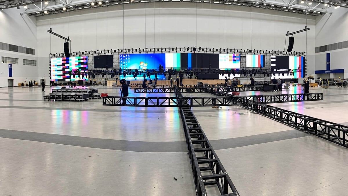 A hall part-way through build: truss sections laid out across the floor, crew working at the stage, and a long run of LED panels showing test patterns.
