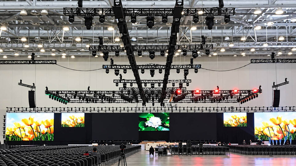 An exhibition hall set for a conference: a row of screens across the stage, moving-head lights and speakers on overhead truss, and rows of banquet chairs facing the stage.