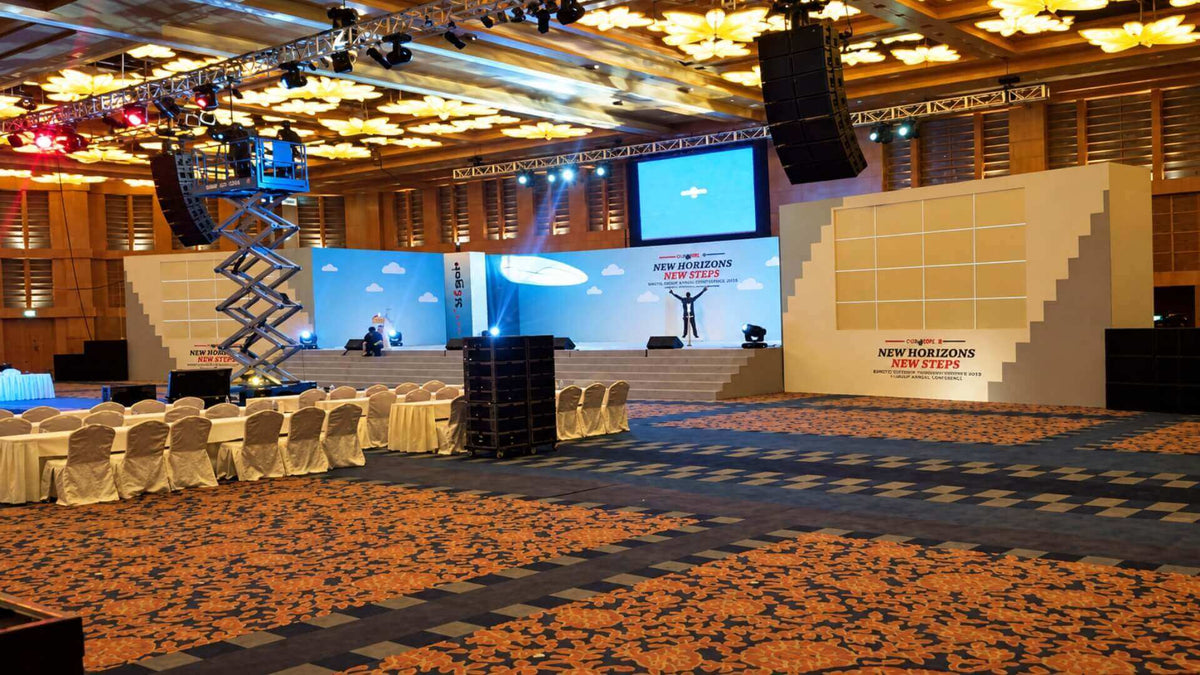 A ballroom being built for a conference: line-array speakers and lighting on truss, a scissor lift at work, and a wide stage set with a projection screen.