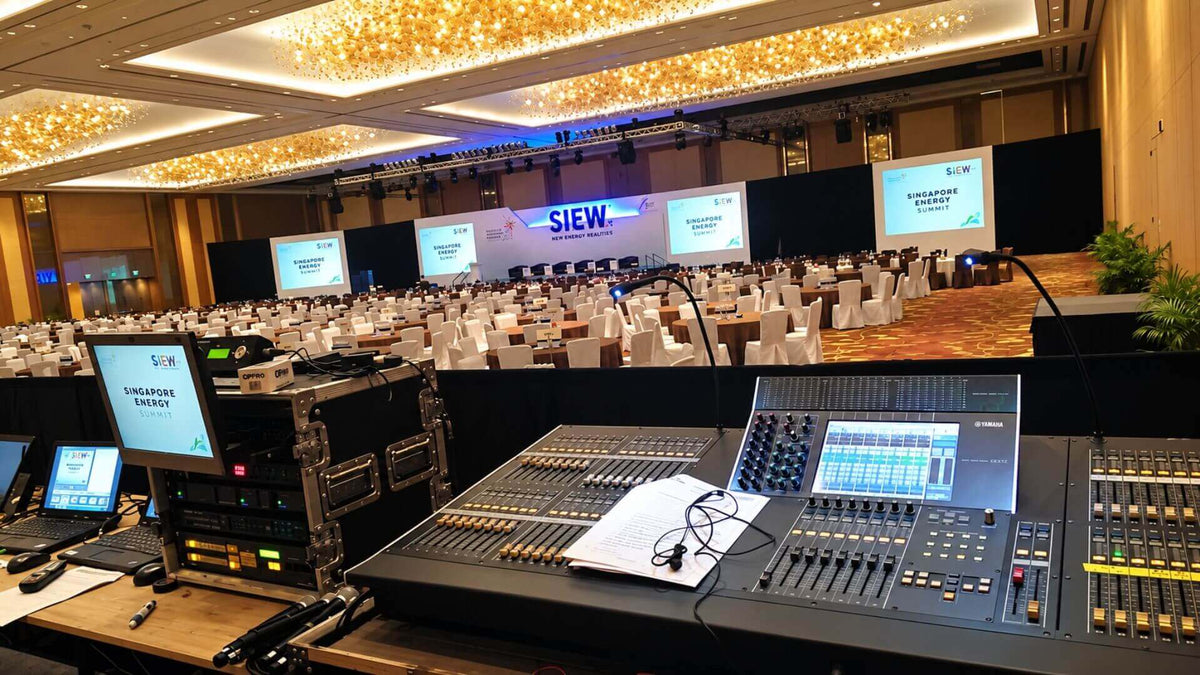The technical control desk at a summit: a mixing console and equipment racks in front of a ballroom of round tables, with four projection screens and a lighting truss over the stage.