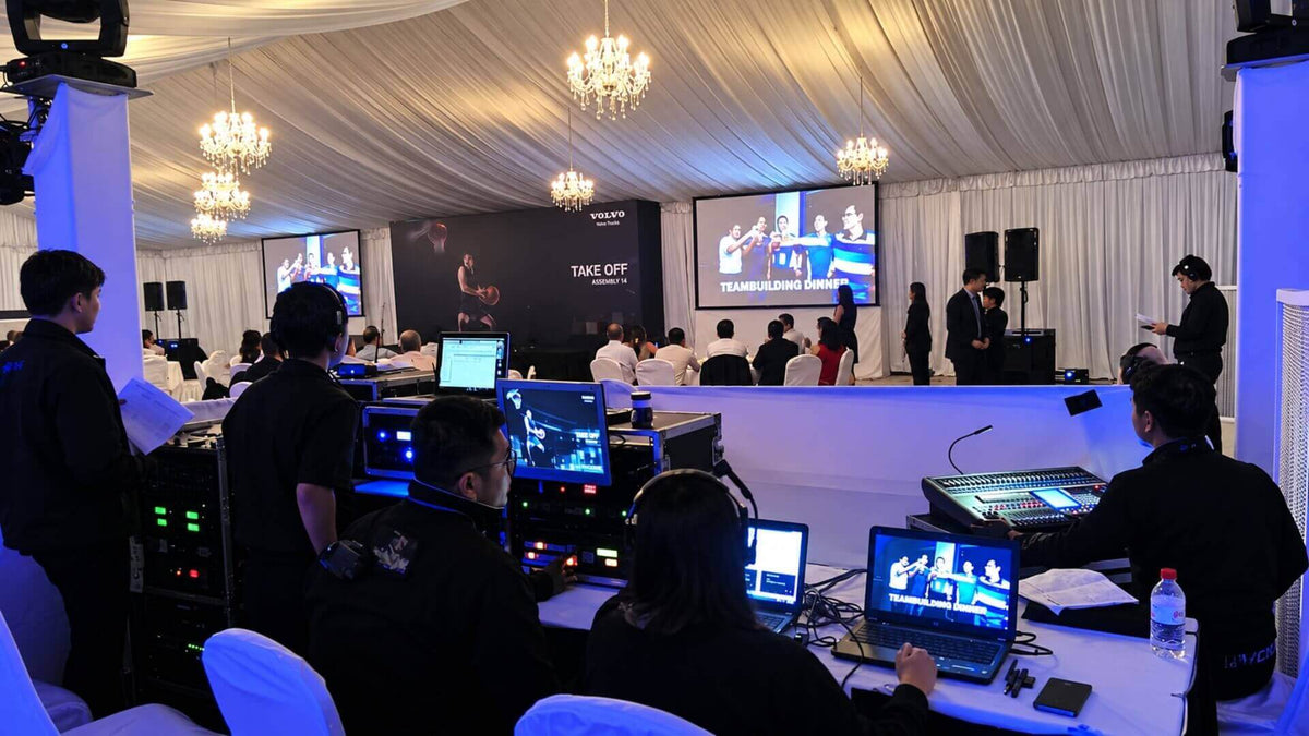 A technical crew at the control desk of a marquee dinner, with a lighting console, laptops and racks in front of two projection screens.