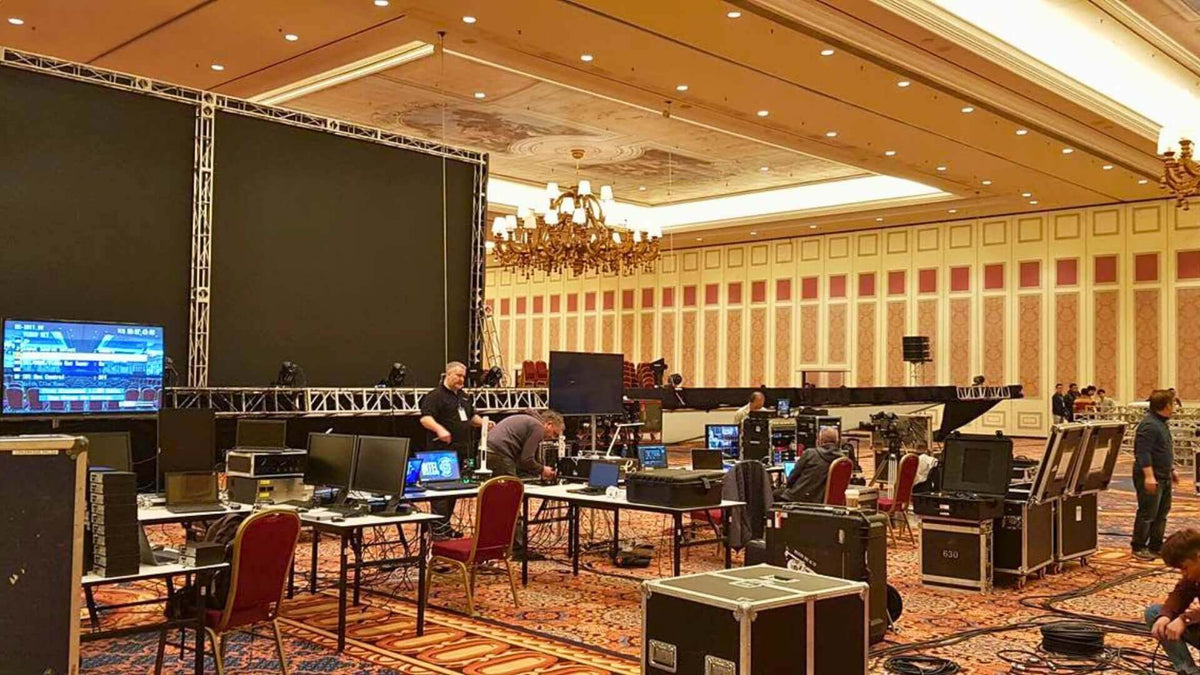 A ballroom during set-up, with a large screen frame on truss, flight cases and technical tables.