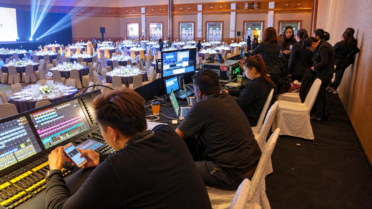 Operators at a lighting console and laptops at the side of a banquet room, with tables set and the stage lit in the background.